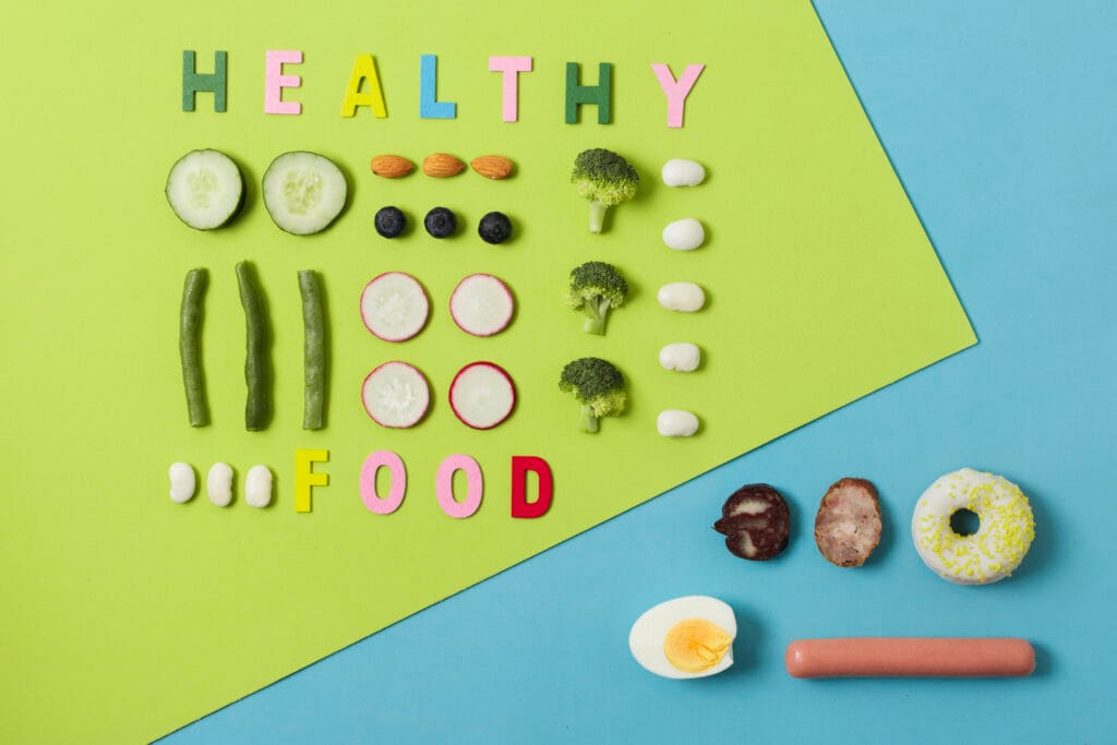 Pieces and slices of healthy food such as beans, cucumbers, egg, broccoli and almonds, are arranged in a decorative structured way on a background of green and blue paper. The words "healthy food" also appear made out of coloured paper