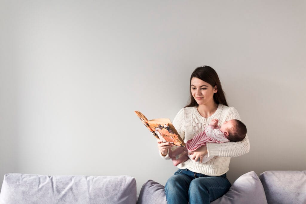 Mother singing a lullaby from book to her baby on her lap in the bedroom