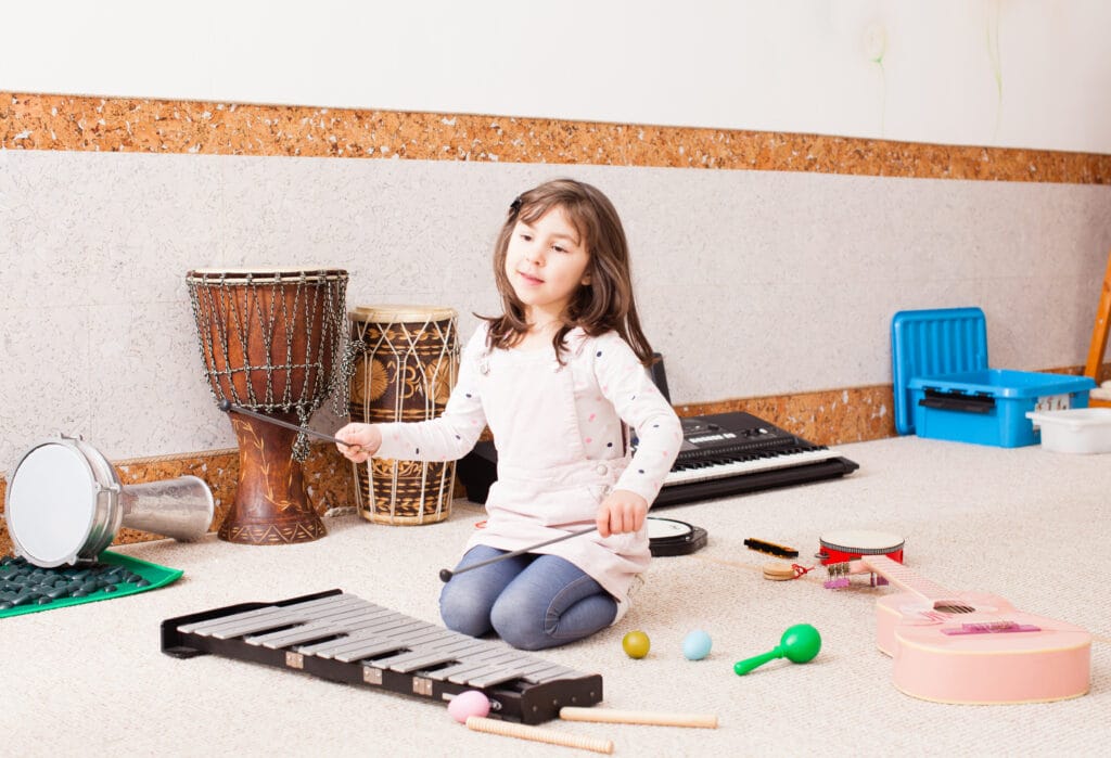 Young girl sits on floor surrounded by various percussion instruments, she is attempting to pay a xylophone