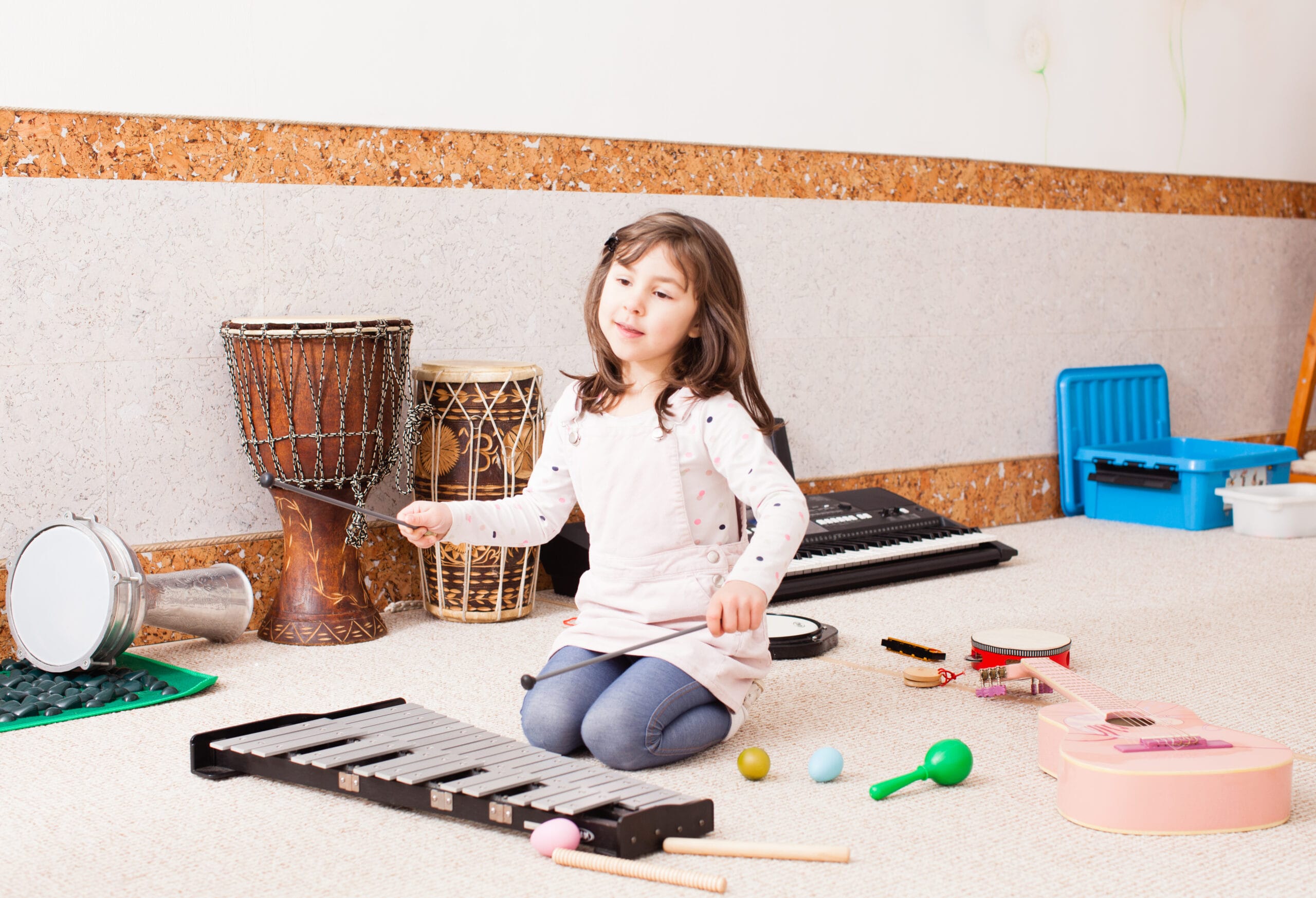 Young girl sits on floor surrounded by various percussion instruments, she is attempting to pay a xylophone