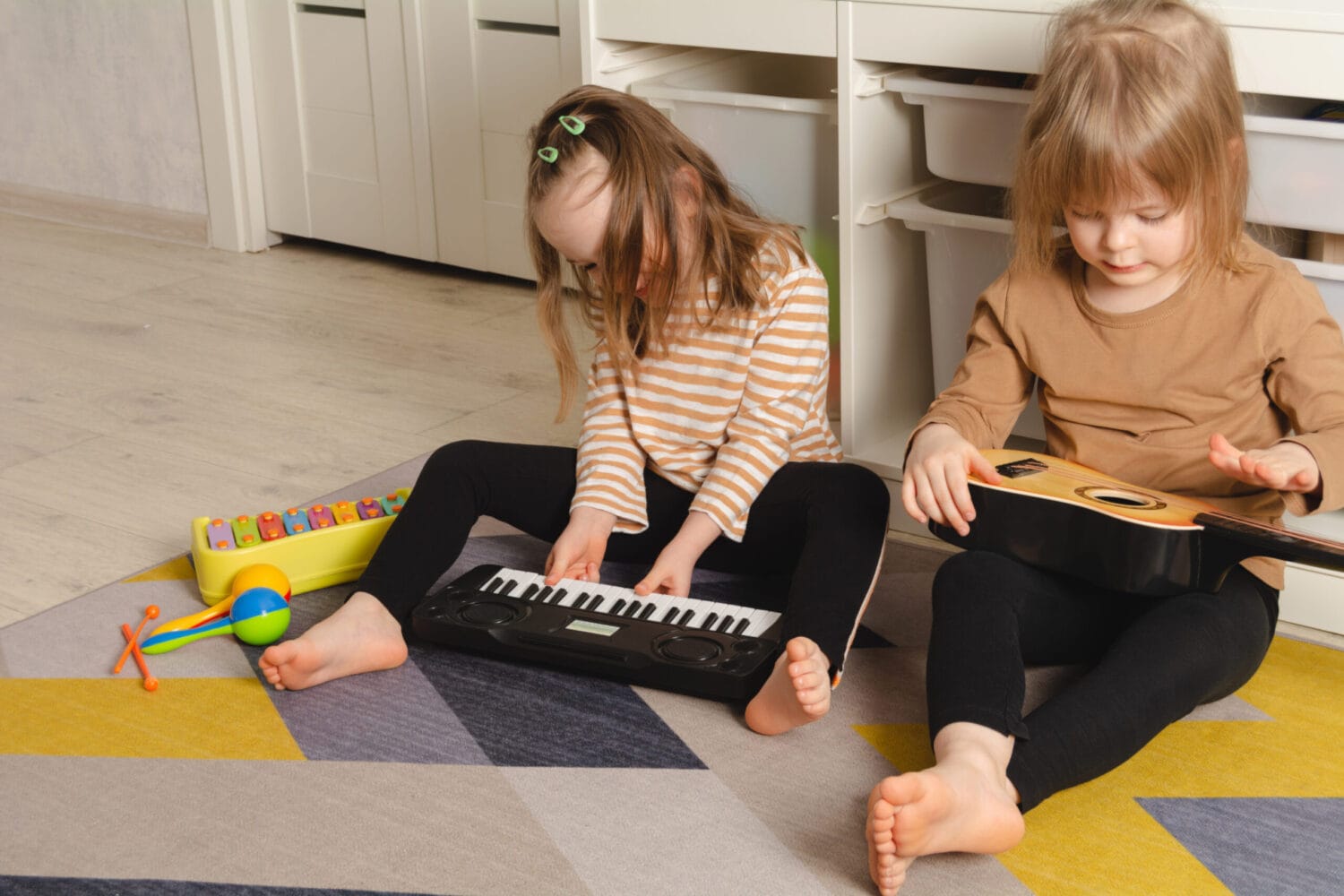 Two young girls sit on the floor of a nursery playing with different musical instruments, one is a keyboard one is a small child sized guitar